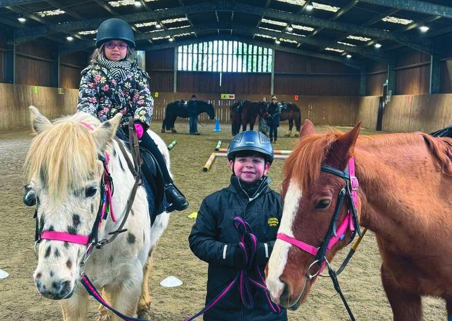 Two children wearing riding hats with a white pony and a chestnut pony in the indoor arena at Cranleigh RDA