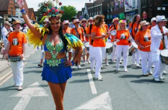 A dancer in a blue and green costume with a feathered headdress leads a Samba band along Cranleigh High Street during the carnival parade