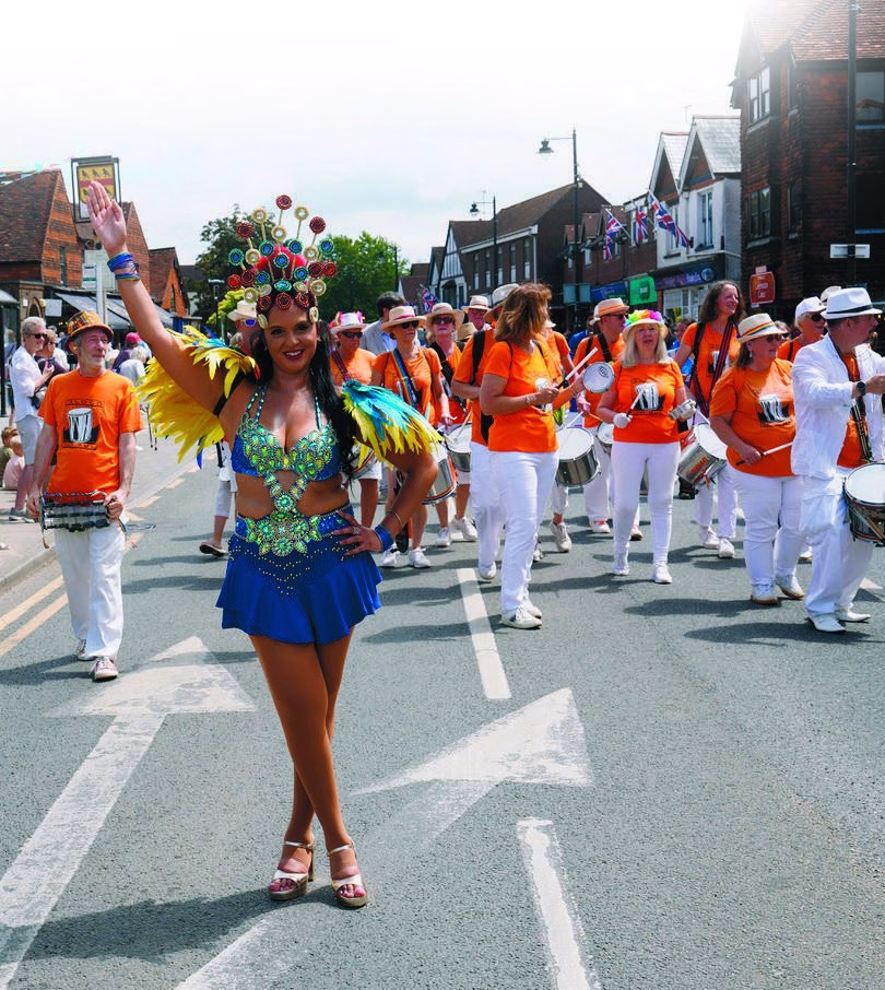 A dancer in a blue and green costume with a feathered headdress leads a Samba band along Cranleigh High Street during the carnival parade