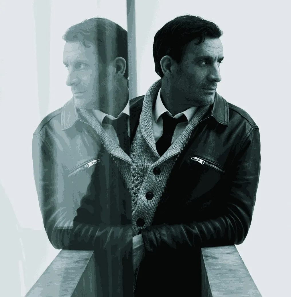Andrew Gow, portrait painter, photographed in a leather jacket beside a reflective window