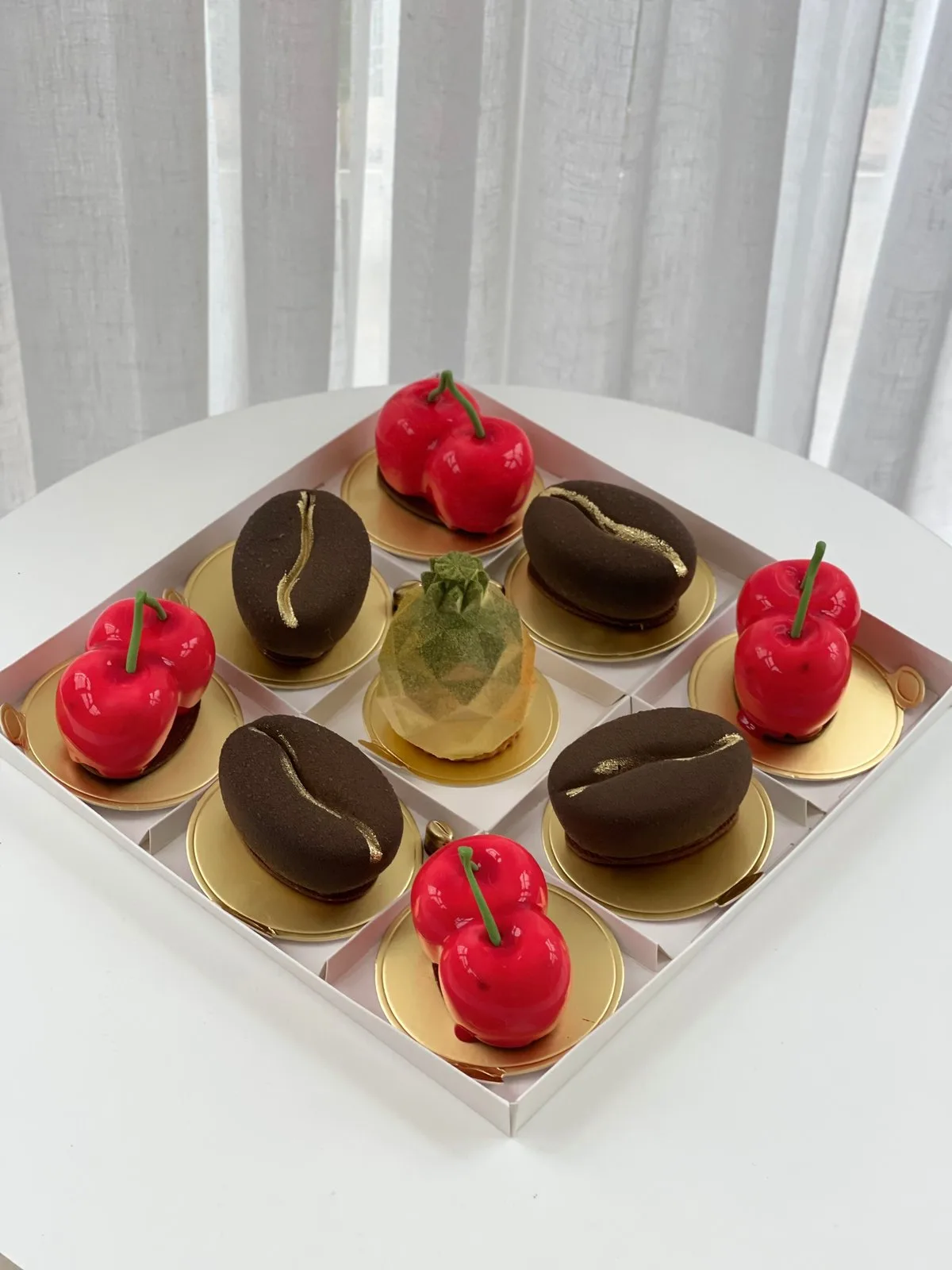 A presentation box of nine individual cakes shaped like red cherries and coffee beans