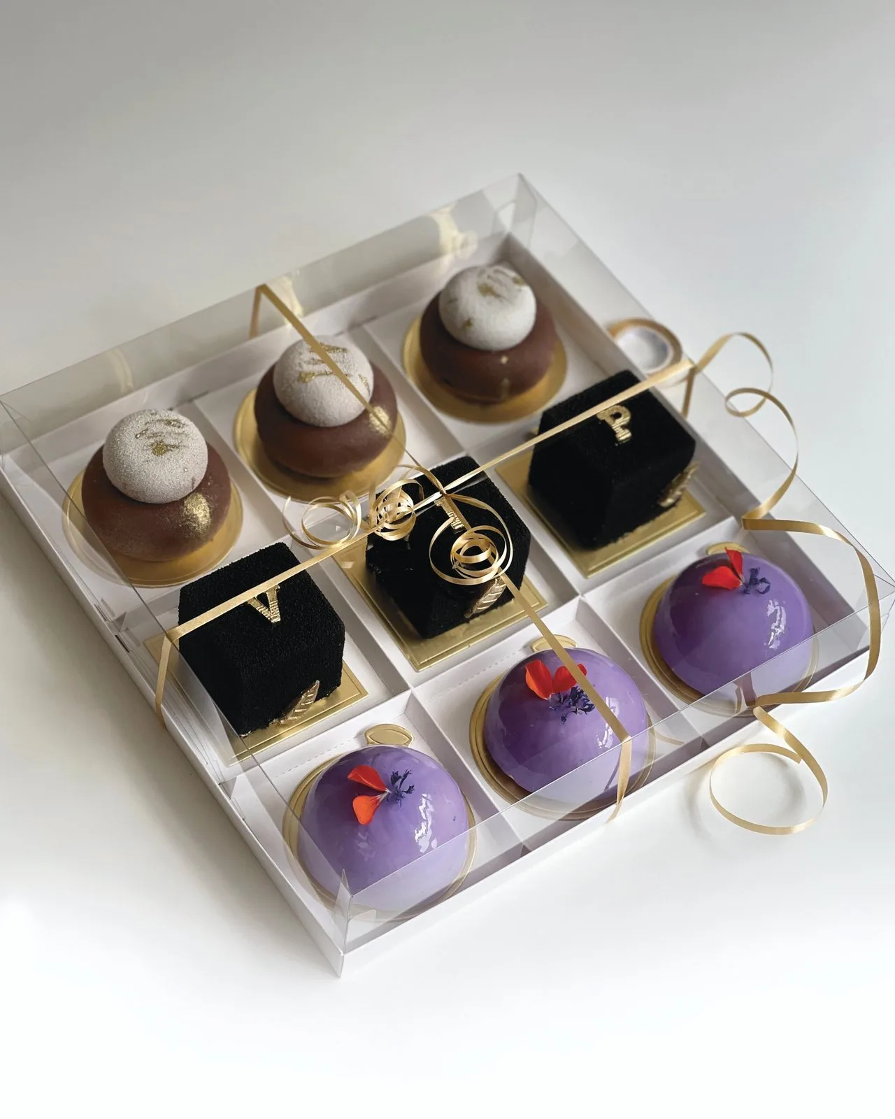 A clear box of nine individual mousse cakes in brown, black and purple, tied with gold ribbon