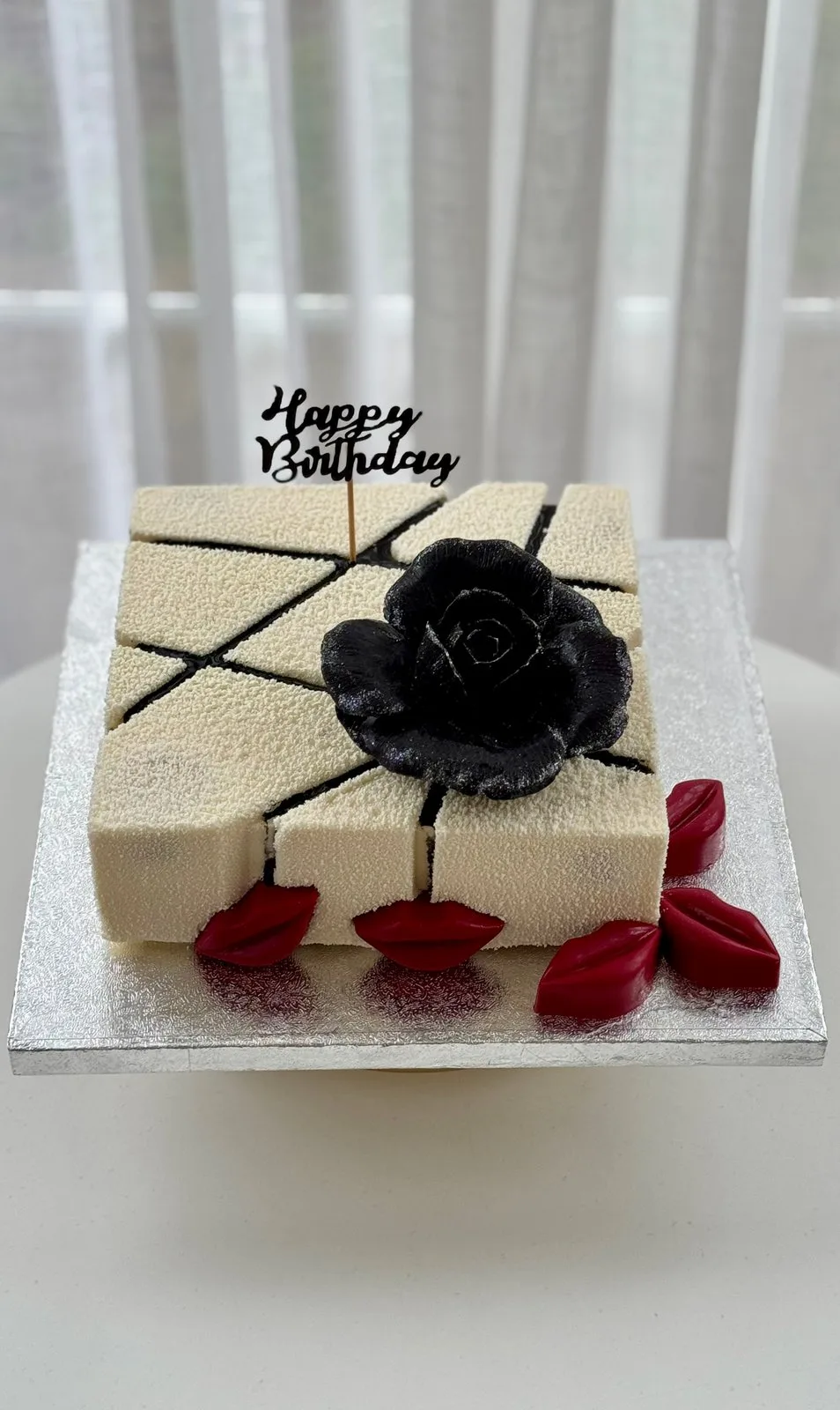 A square cream birthday cake with a black chocolate rose, red lips and a Happy Birthday topper
