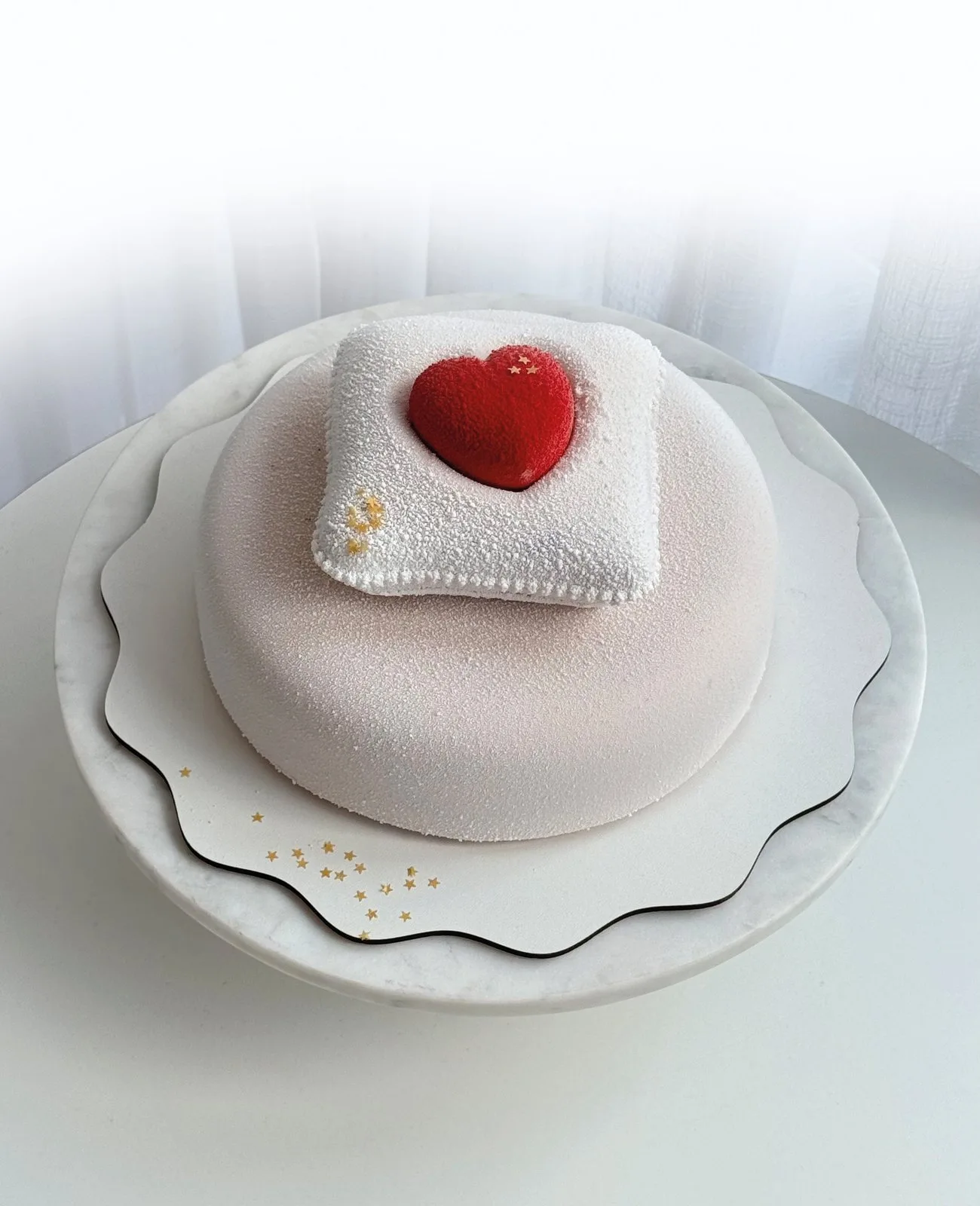 A pale pink cake topped with a small white cushion and a glossy red heart