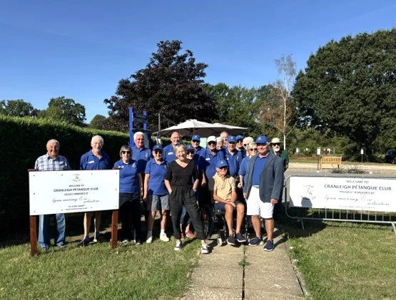 Members of Cranleigh Pétanque Club in blue club shirts, gathered outdoors with Lynn Murray beside two banners